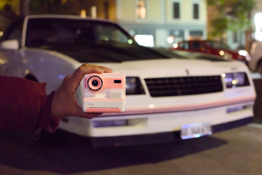 Person holding a pink instant camera in front of a white car at night.