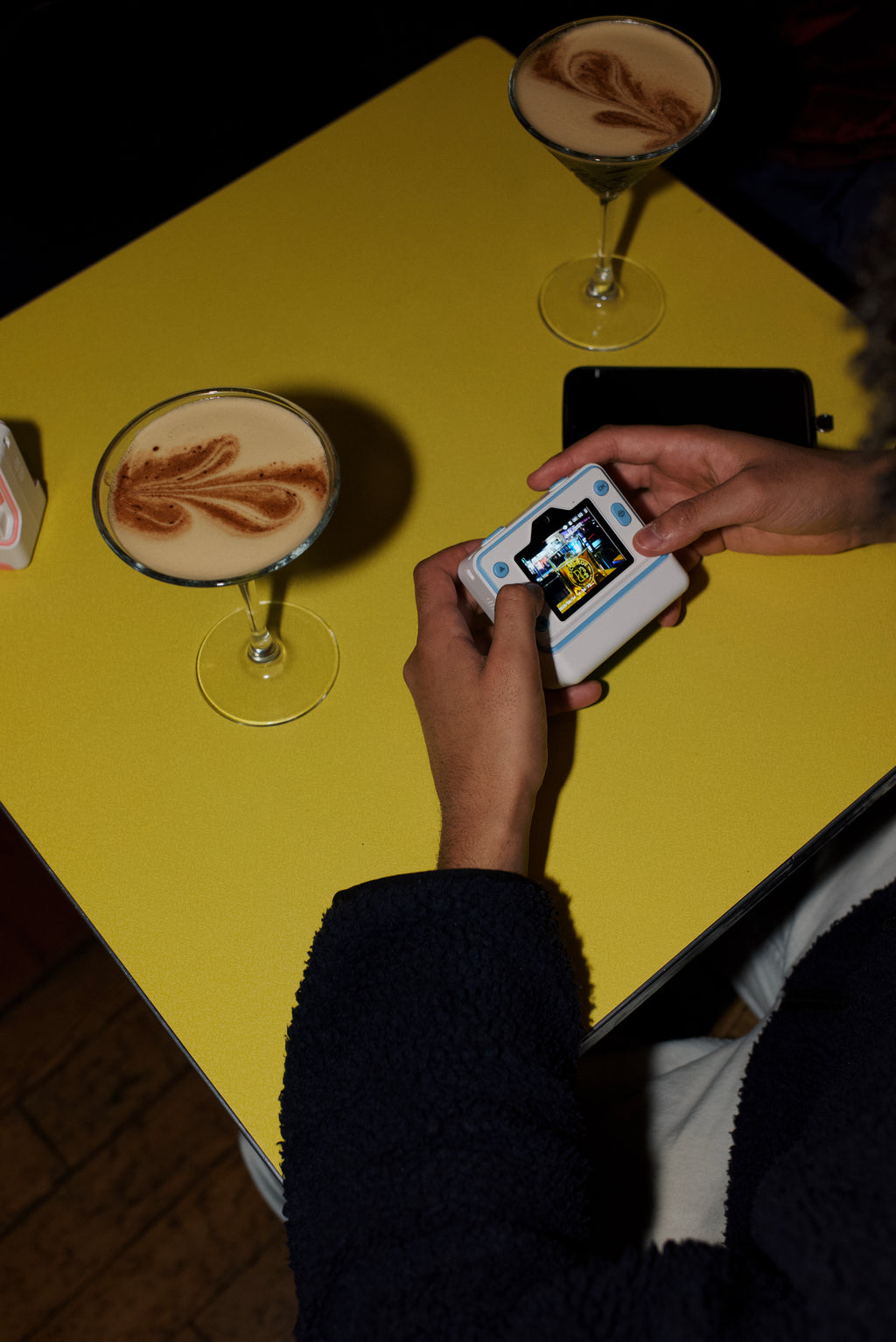 Person using klaq instant camera on a bar table with two cocktails in view.